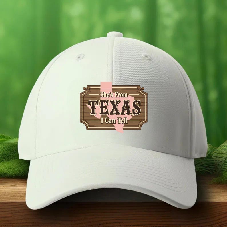 Pu&ograve; includere: Un cappellino da baseball bianco con visiera curva. Il cappellino presenta una grafica marrone e rosa con la scritta "She's From TEXAS I Can Tell". La grafica include un contorno stilizzato dello stato del Texas.