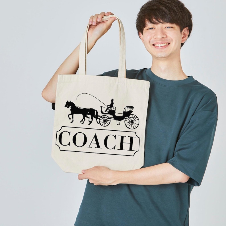 Puede incluir: Un bolso tote beige con un gr&aacute;fico negro de un carruaje tirado por caballos y la palabra "COACH" en un marco rectangular. El bolso lo sostiene una persona que lleva una camiseta verde azulado.