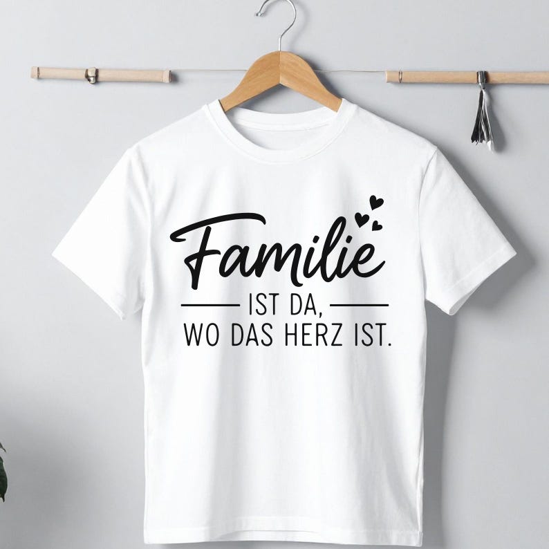Peut inclure: T-shirt blanc avec l'inscription en allemand "Familie ist da, wo das Herz ist" en &eacute;criture noire. Le motif comprend des c&oelig;urs. Le t-shirt est suspendu &agrave; un cintre en bois.