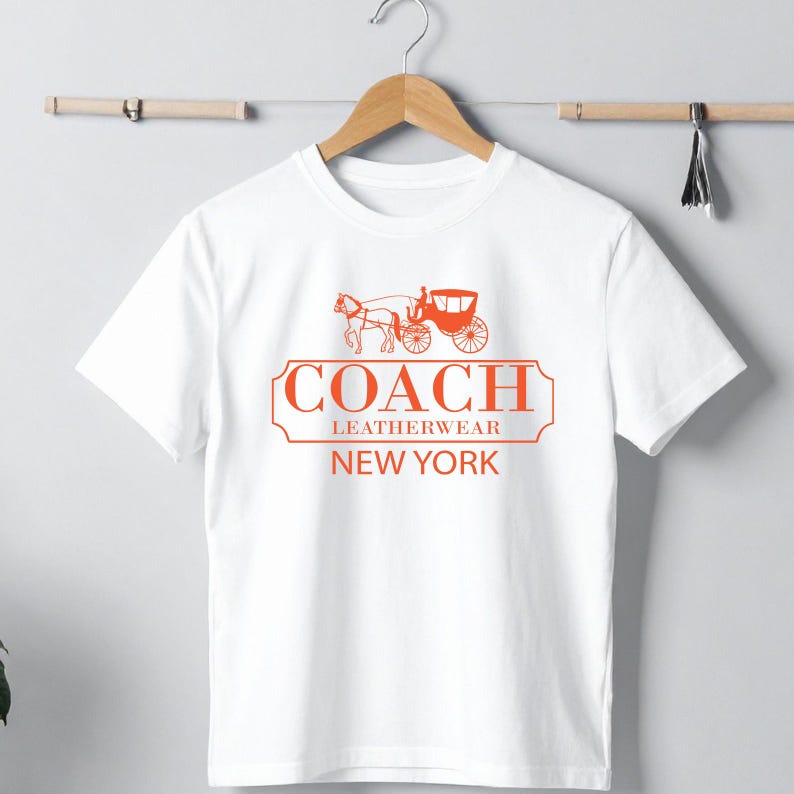 Puede incluir: Camiseta blanca con el logo naranja de Coach Leatherwear. El logo presenta un carruaje tirado por caballos y el texto "COACH LEATHERWEAR NEW YORK". La camiseta est&aacute; colgada en una percha de madera.