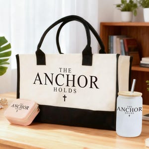 May include: A cream-colored tote bag with black handles and a black bottom panel. The text "THE ANCHOR HOLDS" is printed in black. A small pink square case and a white glass with a straw also display the same text.