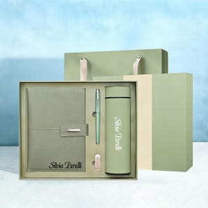 Peut inclure: Un coffret cadeau vert sauge comprenant un carnet, un stylo, un gobelet et un petit clip, le tout présenté dans une boîte. Le carnet et le gobelet portent l'inscription "Silvia Perelli". L'ensemble est présenté avec un sac cadeau assorti.