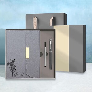 May include: A gray gift set featuring a notebook, pen, and refill, presented in a box. The notebook has a floral design and the name "Donalda" printed on it. The pen is silver and black. The box is gray with a light beige accent.