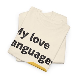 May include: A folded cream-colored t-shirt featuring the black text "My love languages" printed on the front, with a partial yellow graphic visible at the bottom edge of the fabric. The garment is made of a light-colored textile with a subtle speckled texture.
