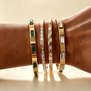 May include: Four gold-toned bangles worn on a wrist. The first features rectangular green stones, the second has a row of small clear baguette stones, the third displays round clear stones, and the fourth has rectangular clear stones.