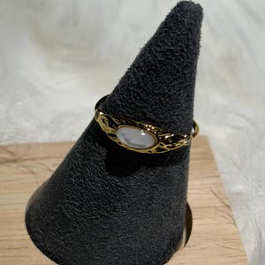 May include: A gold-colored ring with an oval, white stone set in a textured band. The ring is displayed on a black, cone-shaped stand, which is set on a small, light-colored wooden base. The background is a soft, white, fluffy material.