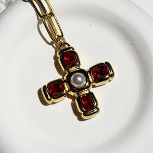 May include: A gold-toned cross pendant necklace with red rectangular accents and a central pearl. The cross is suspended from a gold chain. The necklace is displayed on a white surface.
