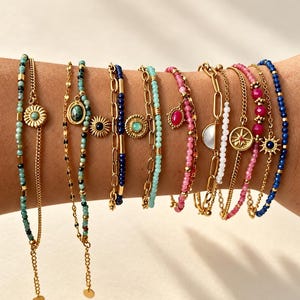 May include: A collection of ten gold-tone chain and beaded bracelets featuring various charms, including sunbursts, oval stones, and geometric links. The bracelets incorporate blue, green, pink, and white beads, with adjustable chain lengths.