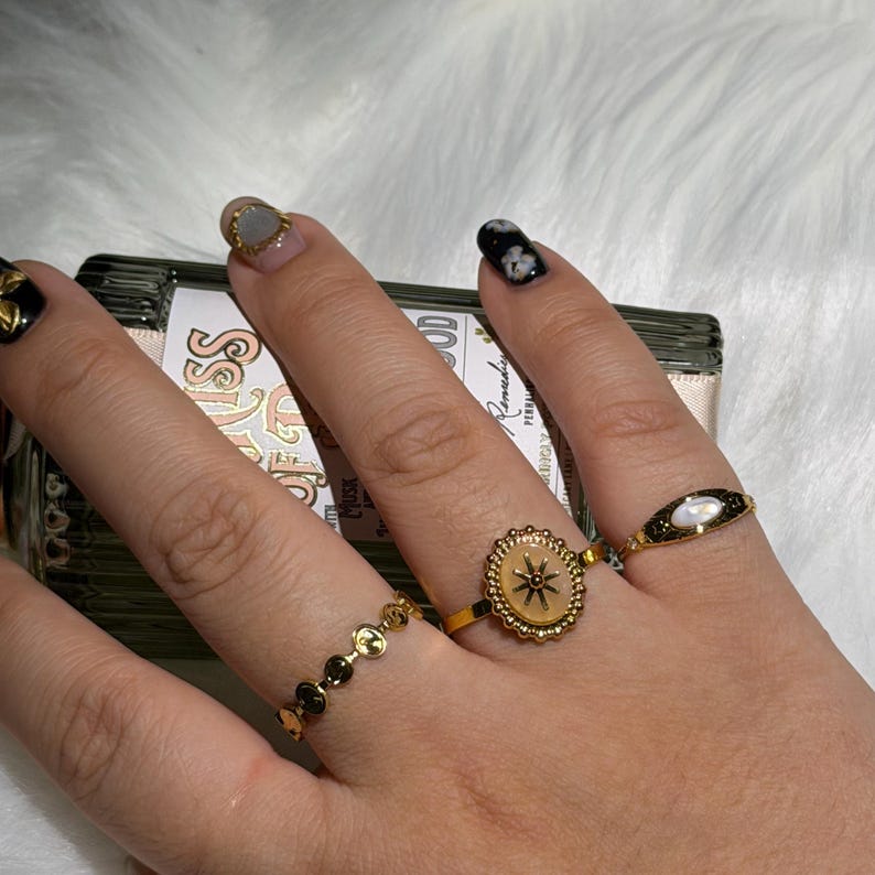 May include: A hand wearing four gold rings. One ring features a starburst design, another has a series of small circles, and a third has a black and white design. The rings are on a hand with manicured nails.