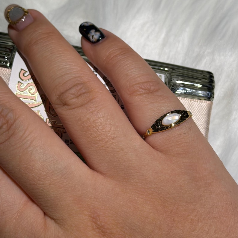 May include: A gold-colored ring with a black enamel design and a central oval-shaped pearl. The ring is on a finger, with a blurred background. The ring has a delicate, vintage aesthetic.
