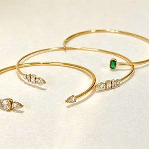 May include: Three gold-toned open cuff bracelets. Two bracelets feature clusters of clear, pear-shaped and rectangular crystals at the ends. The third bracelet features a single rectangular green crystal at one end and a clear crystal at the other.