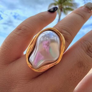 May include: A gold-toned ring featuring a large, irregularly shaped pearl. The pearl has a lustrous, iridescent surface with hints of pink and green. The ring's setting has a textured, organic design.
