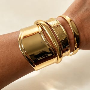 May include: A set of four gold-toned bracelets worn together. The collection includes a wide, smooth cuff, a thick rounded bangle, a medium-width band with a hammered, textured finish, and a thin, polished, smooth circular bangle.