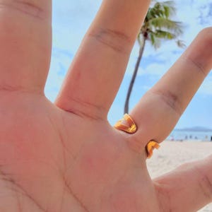 May include: A gold-colored ring with a unique open design, worn on a finger. The ring's shape is angular and modern. The background shows a beach scene with a palm tree and blue sky.