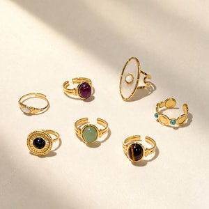 Peut inclure: Un ensemble de sept bagues ajustables dorées ornées de diverses pierres précieuses, dont une grande pierre ovale blanche, un cabochon violet foncé, une pierre noire circulaire avec un bord rayonnant, une pierre verte, un œil-de-tigre et des détails turquoise.