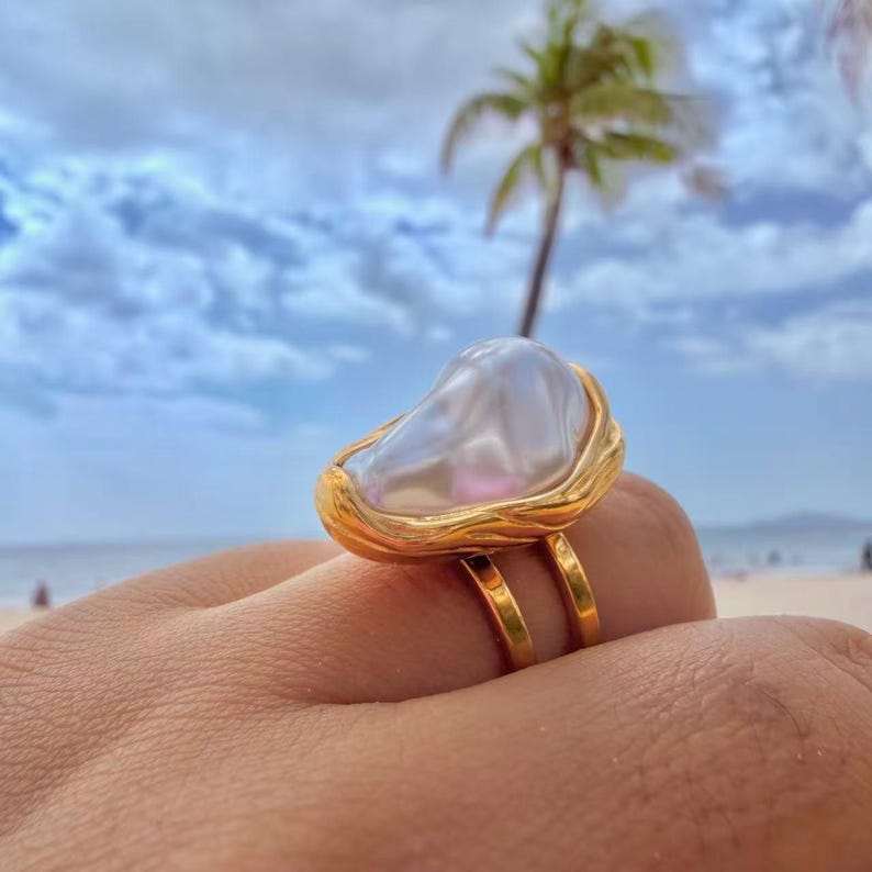 May include: A gold ring featuring a large, irregularly shaped white pearl. The pearl is set in a textured gold bezel, with a double-banded gold ring. The background shows a beach scene with a palm tree and blue sky.