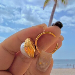 May include: Gold-toned jewelry set featuring a ring and a pair of earrings. The earrings have a pearl and gold design. The ring is a simple gold band. The background shows a blue sky and palm tree.