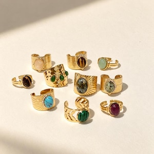 Peut inclure: Une collection de dix bagues en métal doré ornées de diverses pierres précieuses ovales, notamment de l'œil de tigre, de la turquoise, de la malachite et des pierres tachetées. Les bagues présentent des designs variés, comme des textures striées, des motifs rayonnants et des feuilles, avec des structures ouvertes réglables.