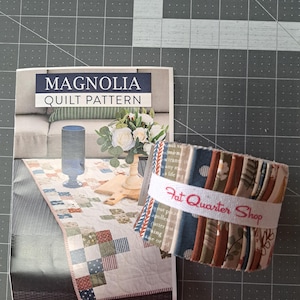 May include: A quilt pattern titled "Magnolia" with a picture of a quilt on a table. A bundle of fabric strips is wrapped with a white band that reads "Fat Quarter Shop". The quilt pattern features a variety of colors.