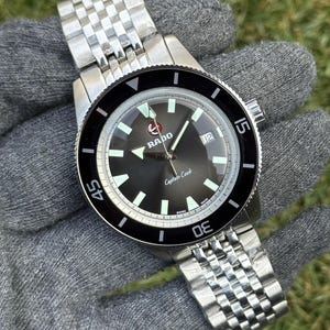 Peut inclure: Une montre Rado Captain Cook en acier inoxydable avec un cadran noir et une lunette rotative noire. La montre présente des marqueurs et des aiguilles luminescents blancs et un guichet de date à 3 heures. Le bracelet est en acier inoxydable.