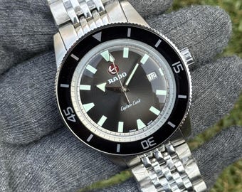 May include: A stainless steel Rado Captain Cook watch with a black dial and a black rotating bezel. The watch features white luminous markers and hands, and a date window at the 3 o'clock position. The watch has a stainless steel bracelet.