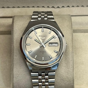 May include: A silver Seiko 5 automatic watch with a silver dial, day and date display, and a stainless steel band. The watch face features the Seiko 5 logo and the text "AUTOMATIC 17 JEWELS".