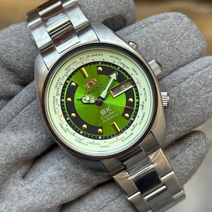 Può includere: Un orologio Orient SK Crystal color argento con quadrante verde e cinturino in acciaio inossidabile. Il quadrante mostra il giorno e la data, oltre alle città del mondo. L'orologio è tenuto su uno sfondo grigio.