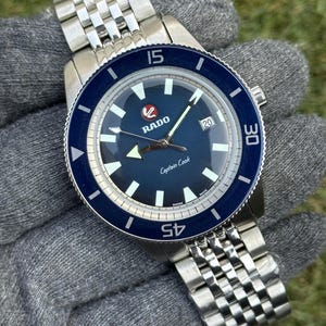 Può includere: Un orologio Rado Captain Cook in acciaio inossidabile con quadrante e lunetta blu. L'orologio presenta un datario a ore 3 e un bracciale in acciaio inossidabile. L'orologio è mostrato su una superficie grigia.
