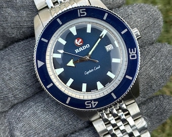 May include: A stainless steel Rado Captain Cook watch with a blue dial and bezel. The watch features a date window at the 3 o'clock position and a stainless steel bracelet. The watch is shown on a gray surface.