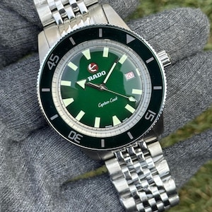 Puede incluir: Un reloj Rado Captain Cook de acero inoxidable con esfera verde y bisel negro. El reloj presenta agujas e indicadores luminosos, una ventana de fecha y un brazalete de acero inoxidable. La esfera del reloj tiene el texto "Captain Cook".