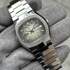 Puede incluir: Un reloj de pulsera automático Mido Multi Star plateado. El reloj tiene una esfera cuadrada con una esfera gris claro, visualización de día y fecha, y agujas e indicadores plateados. La correa del reloj está hecha de eslabones metálicos plateados.