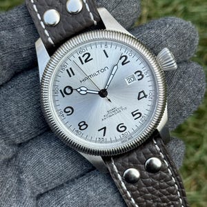 Può includere: Un orologio Hamilton Khaki Automatic argentato con cinturino in pelle marrone. Il quadrante dell'orologio presenta numeri e lancette neri, con un'indicazione della data alle ore 3. Il cinturino è rifinito con cuciture bianche e rivetti argentati.