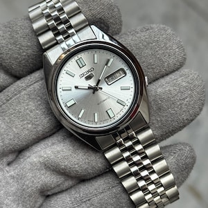 Puede incluir: Un reloj automático Seiko 5 plateado con esfera plateada y correa de acero inoxidable. La esfera muestra el día y la fecha. El reloj se muestra sobre una superficie gris.