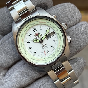 May include: Silver Orient AAA Crystal 21-jewel watch with a white face, featuring a world time bezel and a date display. The watch has a stainless steel strap and a luminous dial with black numerals and hands. The watch is shown on a grey surface.