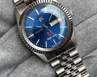 May include: A stainless steel wristwatch with a blue dial. The watch features a fluted bezel, a day-date display, and a silver-tone bracelet. The dial has silver hour markers and the brand name "RADO". The watch is a classic timepiece.