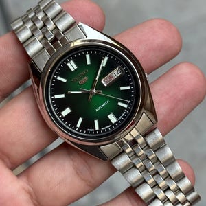 May include: A stainless steel Seiko automatic watch with a green dial. The watch face displays the day and date, with luminous markers and hands. The watch has a silver-tone metal band.