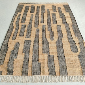May include: A rectangular area rug featuring a woven texture with a repeating pattern of dark charcoal gray vertical stripes against a natural tan background. The rug is finished with short, off-white fringe tassels along the top and bottom edges.