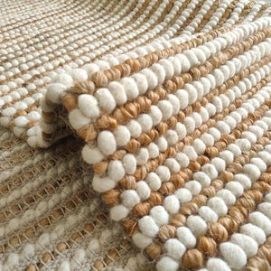 May include: Close-up of a woven rug with a textured surface. The rug features alternating rows of white and tan, creating a striped pattern. The material appears to be natural fibre, with a looped texture adding visual interest.