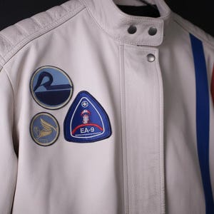 May include: Cream-colored leather jacket with blue and red stripes and patches. The jacket has a stand-up collar, snap closures, and padded shoulders. Patches include a blue circle with a stylized "R", a blue triangle with "EA-9", and a circular patch with a winged design.