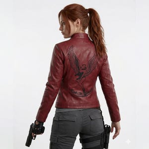 May include: Burgundy leather jacket featuring a black winged figure graphic on the back. The jacket has a classic biker design with a zip-up front and a fitted shape. The text "Made in Heaven" is below the graphic.