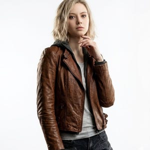 May include: A brown leather jacket with a hooded gray lining. The jacket has a classic biker style with a notched collar, asymmetrical zipper, and multiple pockets. The model is wearing a gray t-shirt and dark jeans.