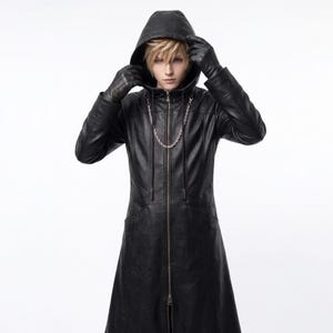 May include: A long black hooded coat featuring a full-length front zipper, long sleeves, and a silver-toned chain draped across the chest. The coat is worn with matching black gloves, and the hood is pulled up over a head of light blonde hair.