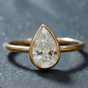 Könnte beinhalten: Ein goldener Ring mit einem einzelnen birnenförmigen, facettierten, klaren Edelstein in einer glatten Zargenfassung. Die Ringschiene ist dünn und poliert und unterstreicht die reflektierende, facettierte Oberfläche des zentralen Steins.