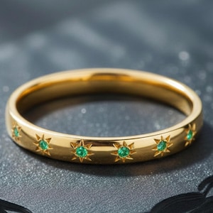 Puede incluir: Un anillo dorado con diseños en forma de estrella, cada uno con una pequeña gema verde. El anillo se encuentra sobre una superficie oscura y texturizada. Una pieza de joyería elegante.