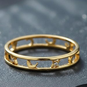May include: A gold-colored ring with a celestial design featuring moons and stars. The ring has a continuous band with cut-out shapes and small, clear gemstones. The ring is displayed on a dark, textured surface.