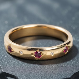 May include: A gold band ring featuring three red gemstones and two smaller clear stones. The ring has a polished finish and is set against a dark grey background. The gemstones are set in star-shaped settings.