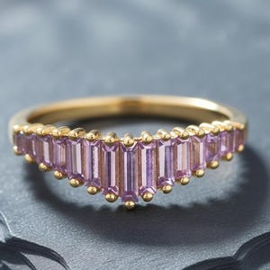 May include: A gold ring featuring a V-shaped chevron band set with a row of light purple baguette-cut stones. The stones are arranged vertically along the front of the band, secured by small gold prongs between each individual gemstone.