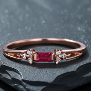 Peut inclure: Bague en or rose ornée d'un rubis rectangulaire au centre, flanqué de petits groupes de pierres blanches. La bague a un anneau fin et des motifs ajourés décoratifs de chaque côté du rubis. La bague est présentée sur une surface sombre et texturée.