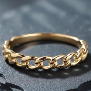 May include: A gold chain link ring. The ring has a continuous pattern of interlocking links, creating a textured surface. The ring is set against a dark, textured background, highlighting its metallic sheen. A piece of jewellery.
