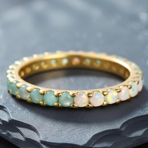 May include: A gold ring with a band of alternating opal and aquamarine gemstones. The ring is set on a dark gray, textured surface. The gemstones are round and closely set, creating a continuous pattern around the ring.